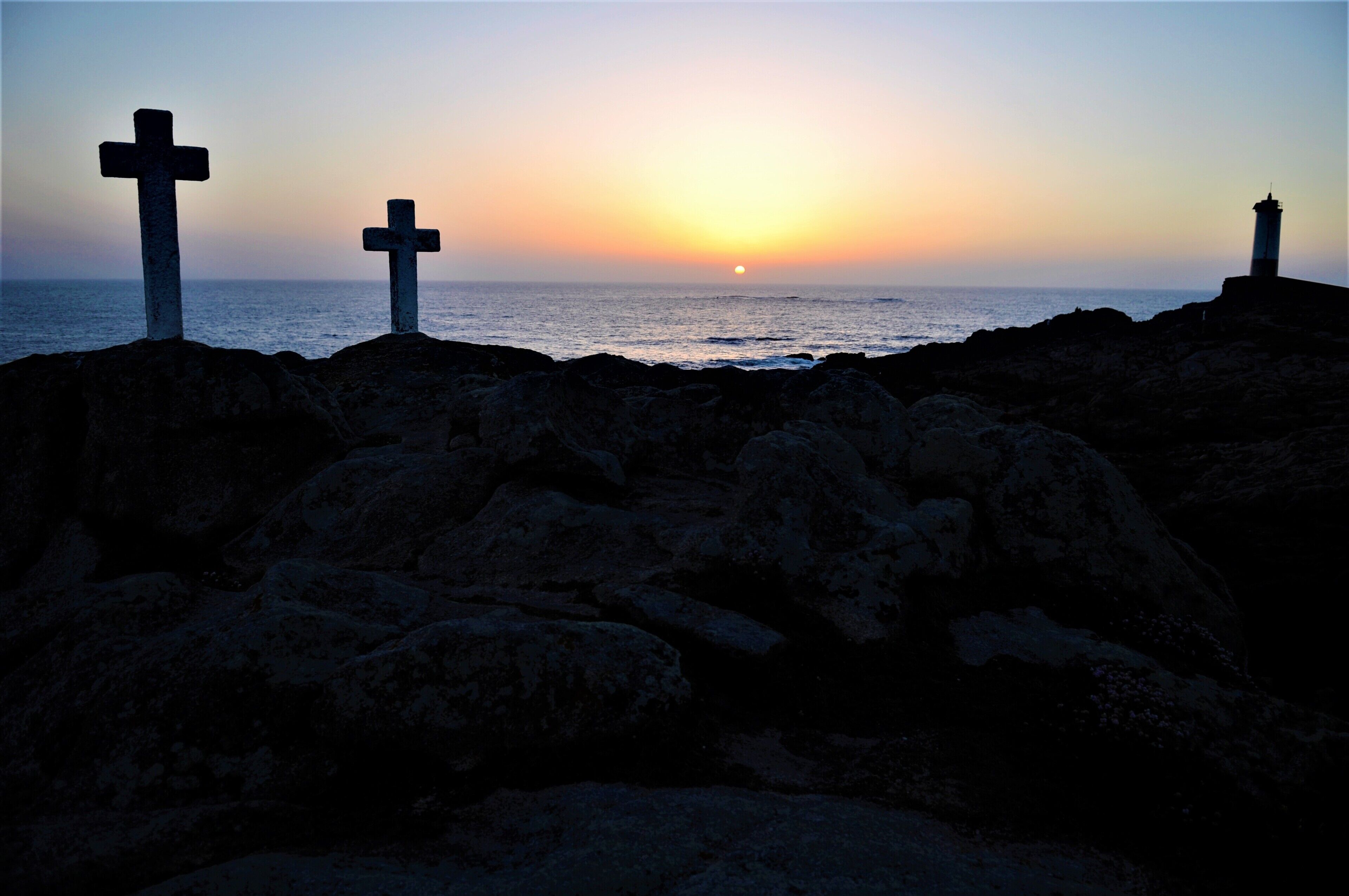 Three crosses on a rocky shoreline with the ocean and a lighthouse in the background.