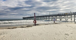 A beach with a wooden pier extending into the ocean, a lifebuoy on the sand, and footprints leading towards the water.