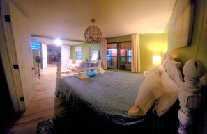A hotel room with a bed, a large stuffed animal, a chandelier, and a view of the hallway.