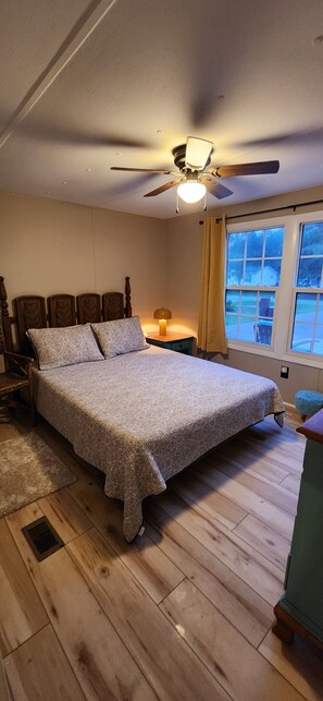 A bedroom with a bed, wooden headboard, a ceiling fan, a window with curtains, and a nightstand with a lamp.