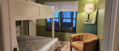 A room with bunk beds, a chair, a window with blinds, and a lamp.