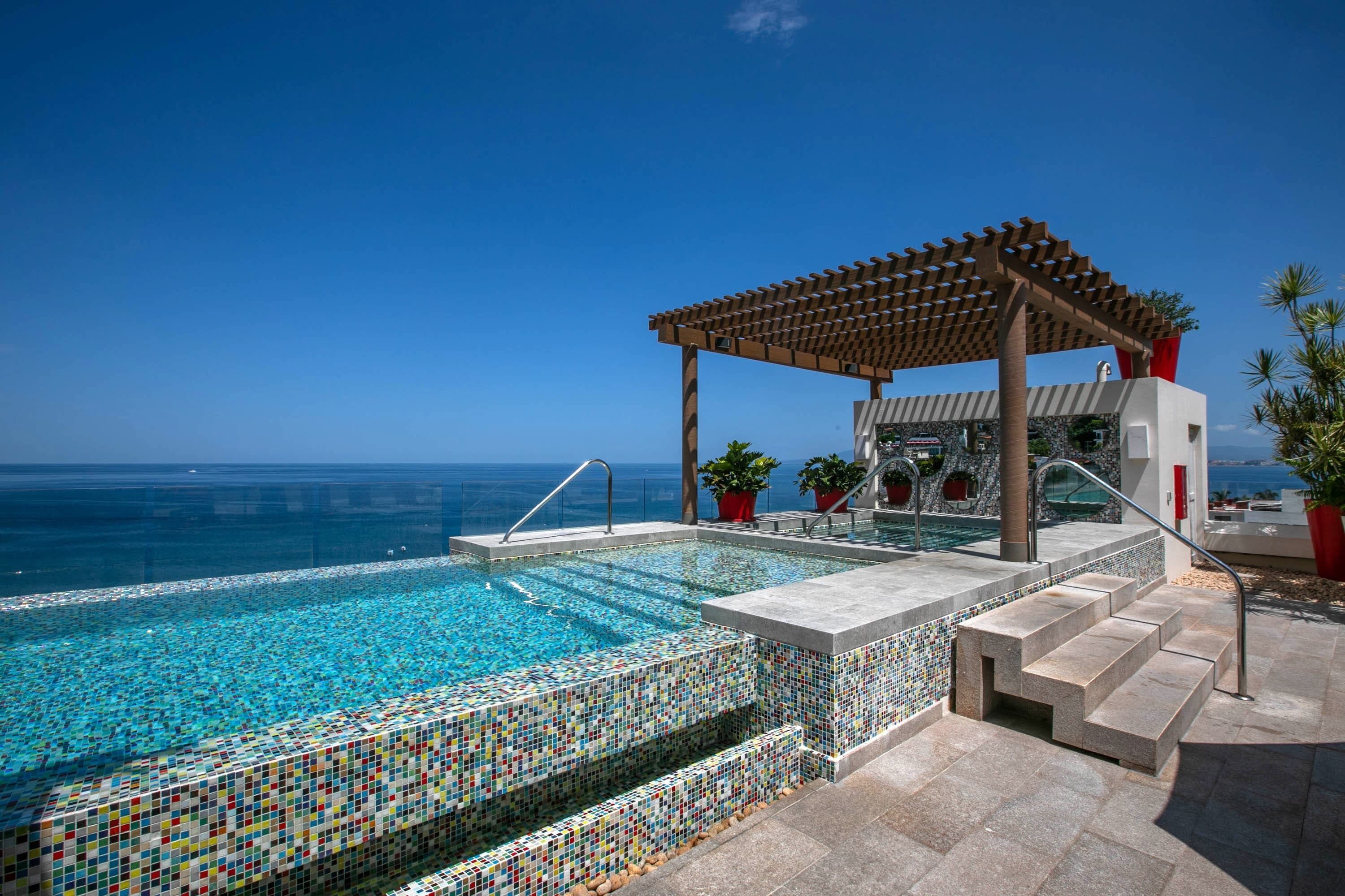 A pool with mosaic tiles, a wooden pergola, and a view of the sea.