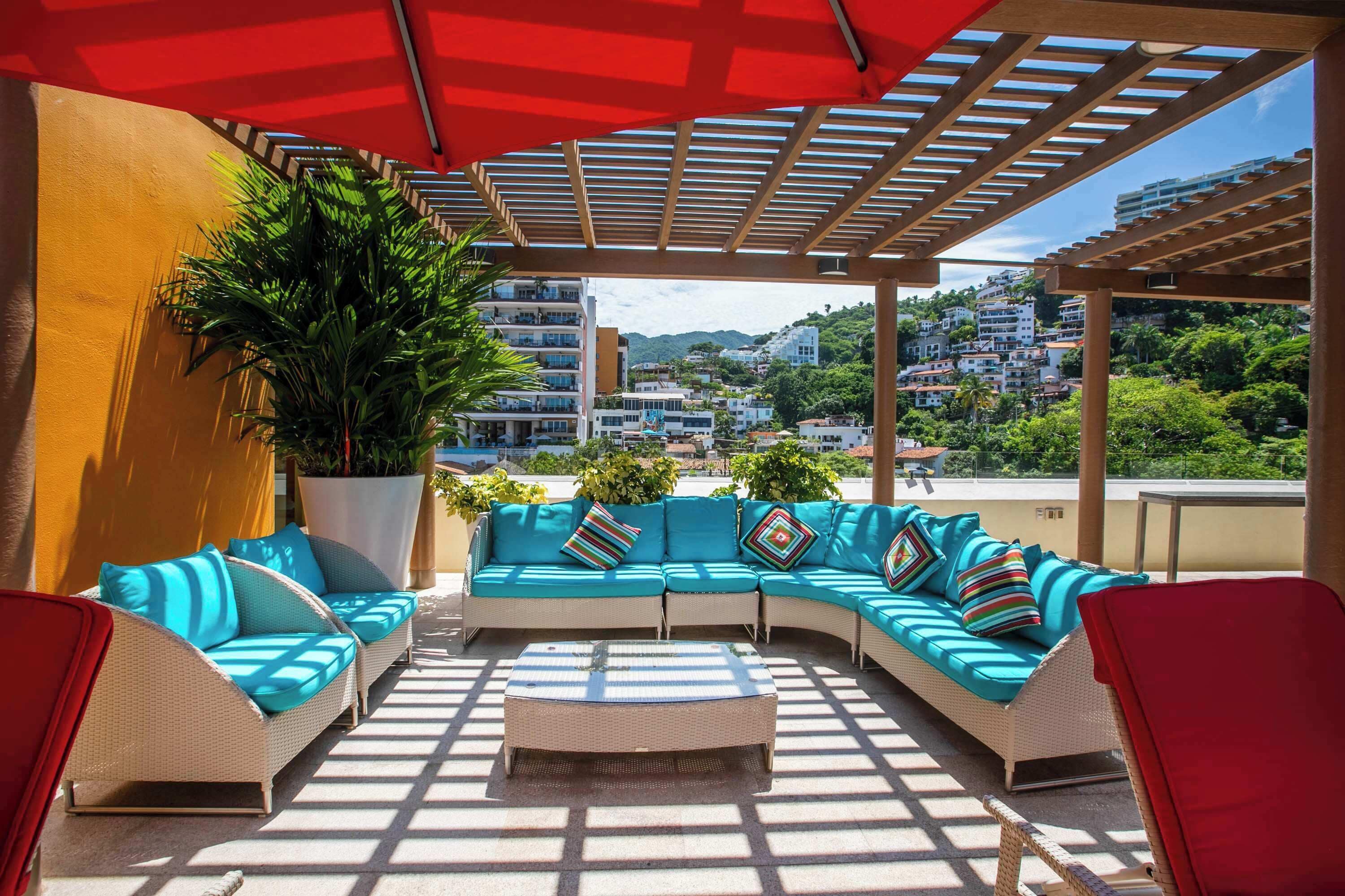 A covered outdoor seating area with a sofa and chairs, a large potted plant, and a view of a hillside town.