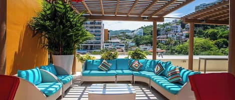 Un espacio de estar al aire libre cubierto, con sofá y sillas, una planta grande en maceta y vistas a un pueblo en la ladera.