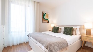 Una habitación de hotel con una cama, una mesita de noche, una lámpara, un cuadro en la pared y cortinas traslúcidas.