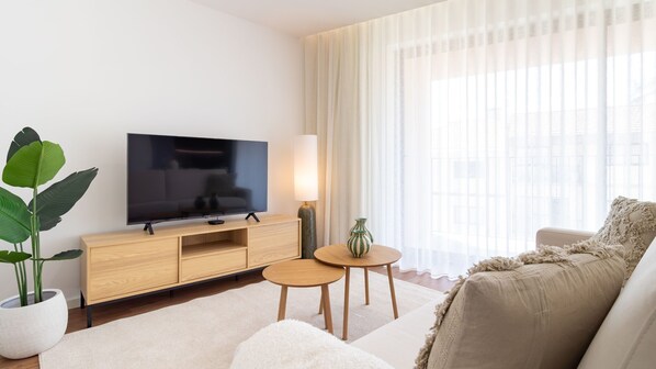 Een moderne woonkamer met een flatscreen televisie, houten meubels, een potplant, een lamp en een groot raam met dunne gordijnen.