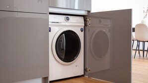 A compact, modern washing machine with a front-loading door, situated in a kitchen area with a wooden floor and a white chair in the background. - Polacos Residence | 2BR | Apt S by LovelyStay (Vila Nova de Gaia)