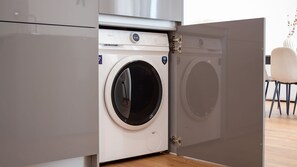 A compact, modern washing machine with a front-loading door, situated in a kitchen area with a wooden floor and a white chair in the background. - Polacos Residence | 2BR | Apt S by LovelyStay (Vila Nova de Gaia)