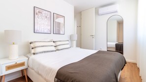 Ein Hotelzimmer mit einem Bett, zwei Nachttischlampen, einem weißen Nachttisch, einer Wand-Klimaanlage und einem großen Spiegel.