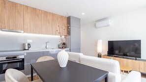 Eine moderne Küche mit einem Backofen von Bosch, einer Spüle und einem Kühlschrank von Bosch. Ein Essbereich mit einem weißen Sofa, einem dunklen Tisch und einer Vase mit Trockenblumen. Ein Flachbildfernseher ist an der Wand montiert und eine Lampe steht auf einem Beistelltisch.