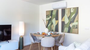 Ett modernt vardagsrum med en platt-TV, en hög golvlampa, en matplats med ett runt bord och två abstrakta tavlor på väggen.