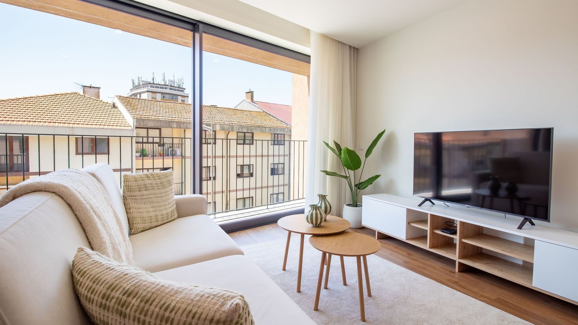 A modern living room with a sofa, TV, and a view of buildings through large windows.