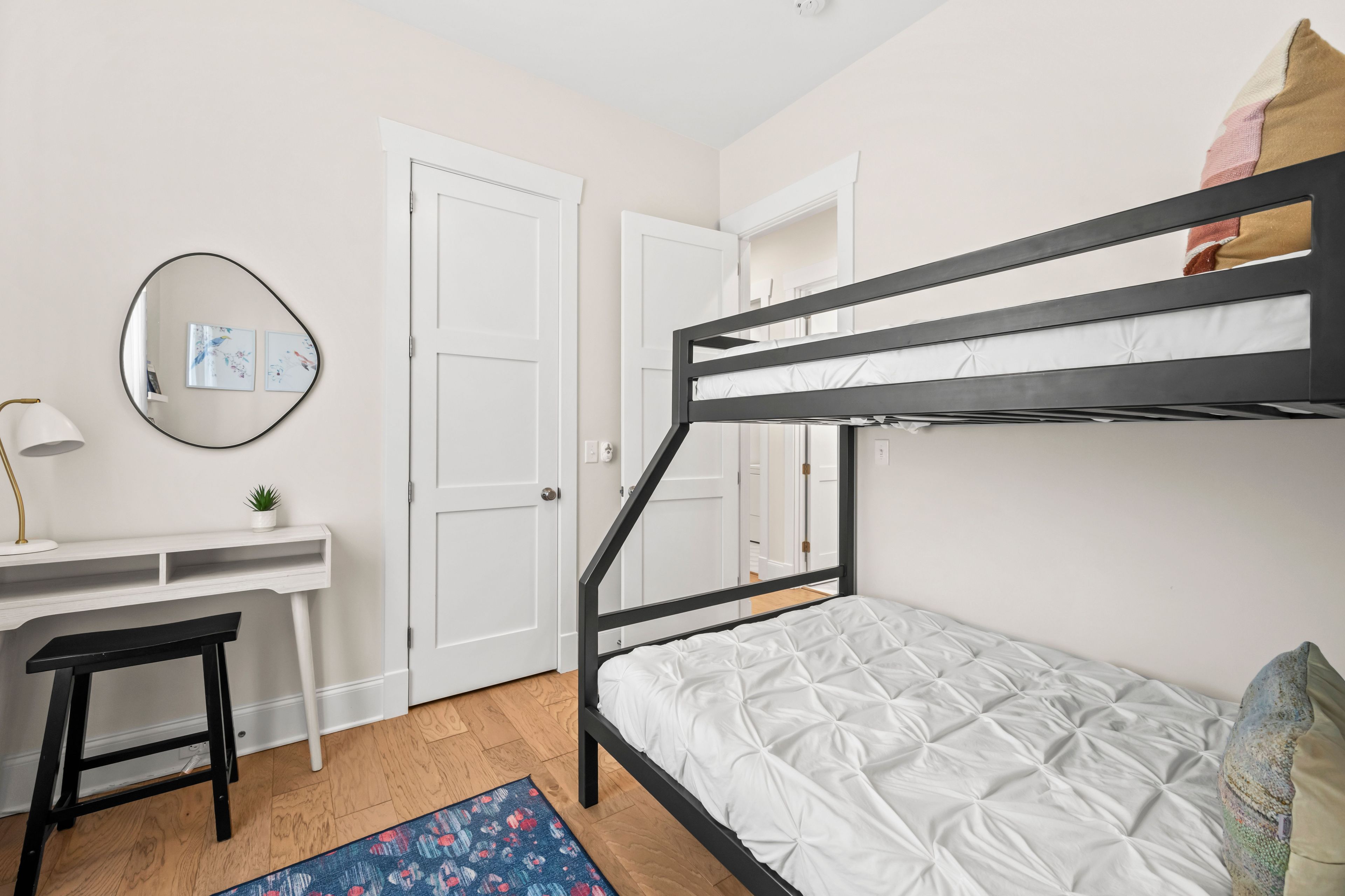 En køjeseng med hvid madras, et skrivebord med lampe, en lille potteplante og et spejl på væggen.