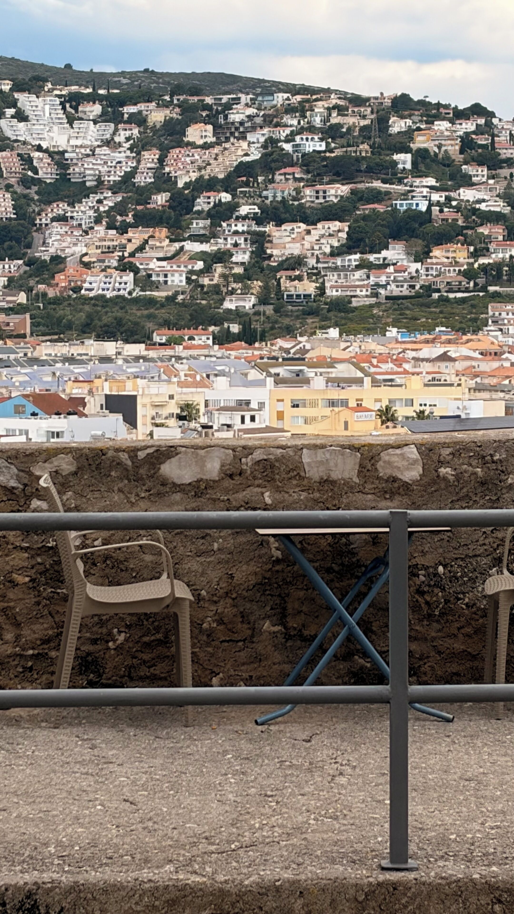 Un paisaje urbano costero con edificios juntos, cielo despejado y primer plano rocoso con barandilla de metal.