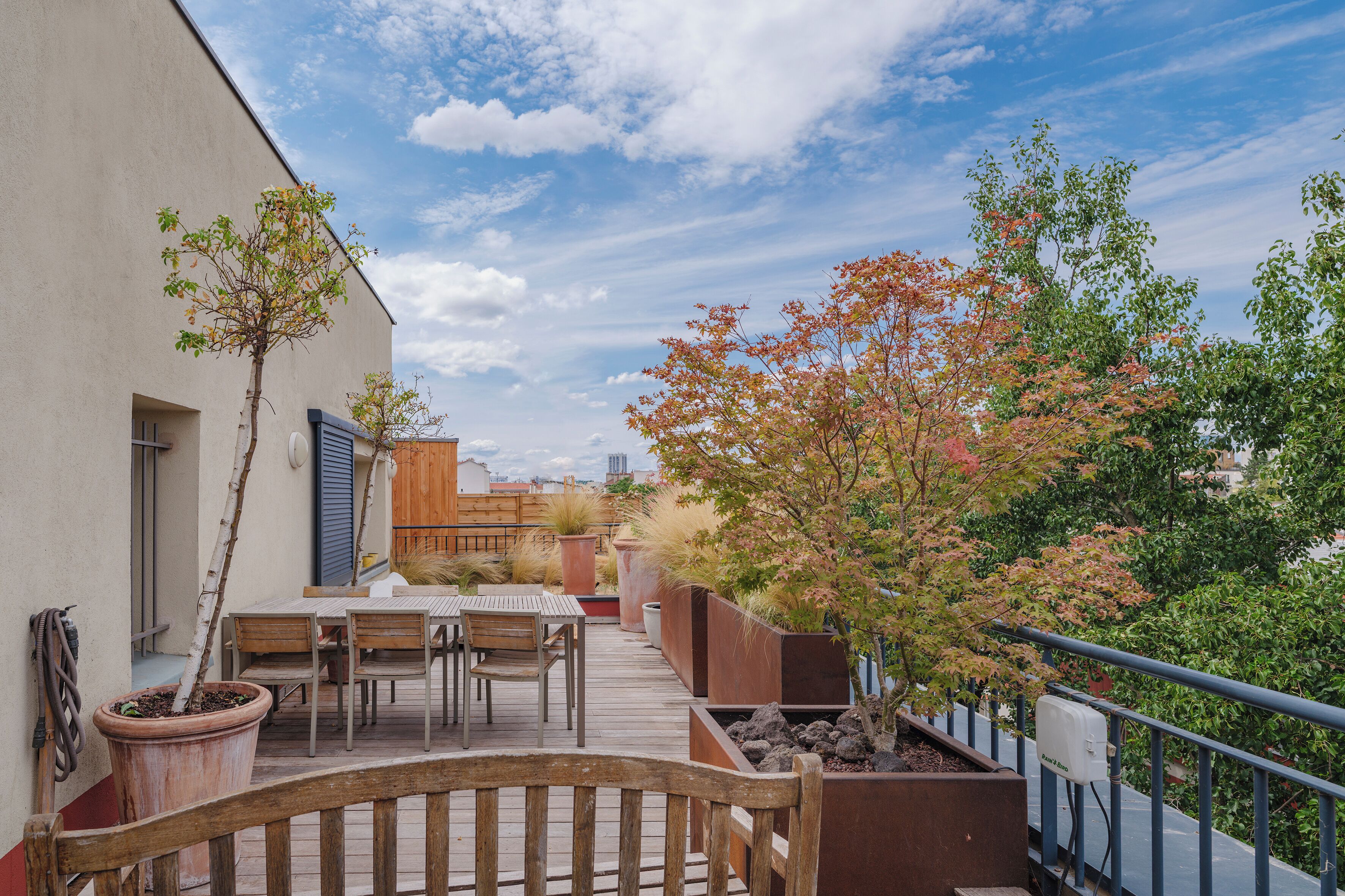 A rooftop terrace with a wooden table and chairs, potted plants, and a view of the cityscape.