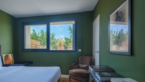 Une chambre à coucher comprenant un lit, une chaise, un bureau avec des livres et deux fenêtres donnant sur un paysage verdoyant.