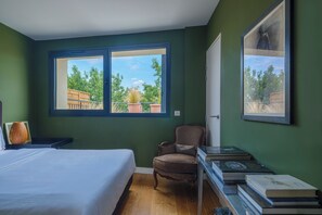 Ein Schlafzimmer mit einem Bett, einem Sessel, einem Schreibtisch mit Büchern und zwei Fenstern mit Blick auf Grünflächen.