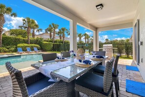 A poolside dining area with a table set for two, surrounded by wicker chairs, overlooking a swimming pool and palm trees. - The Encore Bliss -  Luxury 9-Bed Villa with Oversized Pool & Spa (Kissimmee)