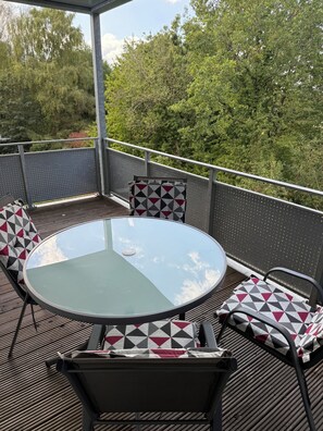 Une table ronde en verre, entourée de quatre chaises, chacune agrémentée d’un coussin à motifs géométriques, est placée sur une terrasse en bois dotée d’une rambarde en métal.