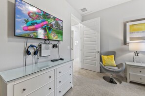 Una sala de estar moderna con un televisor de pantalla plana montado en la pared, una mesa de centro de vidrio, un sillón gris con un cojín amarillo, una mesita de noche blanca con una lámpara y un mueble blanco con cajones.