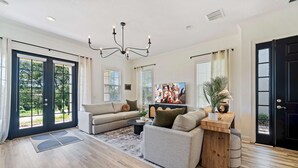 Ein geräumiges Wohnzimmer mit einer großen Couch, einem Couchtisch und einem Fernseher. Große Fenster mit Jalousien, ein Holzboden und eine schwarze Tür sind ebenfalls vorhanden.