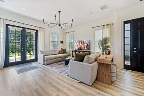 A spacious living room with a large sofa, a coffee table, and a television. There are large windows with blinds, a wooden floor, and a black door. - Summer Breeze - Four Bedroom Villa, Sleeps 8 (Kissimmee)