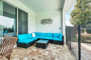 A modern outdoor seating area with a blue sectional sofa, a wooden chair, and a glass table on a paved patio. - Private south facing  Pool and Hot Tub-games room -ping pong-Close to disney (Lake Buena Vista)