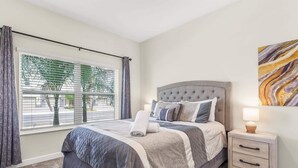 A bedroom with a bed, bedside tables, a window with curtains, and a wall-mounted artwork. - Birdie Bungalow - Four Bedroom House, Sleeps 8 (Kissimmee)