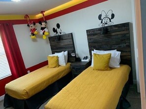 Ein Hotelzimmer mit zwei Einzelbetten, jeweils mit gelber Bettdecke und weißen Kissen. An der Wand befinden sich Minnie Maus und Mickey Mouse Dekorationen.