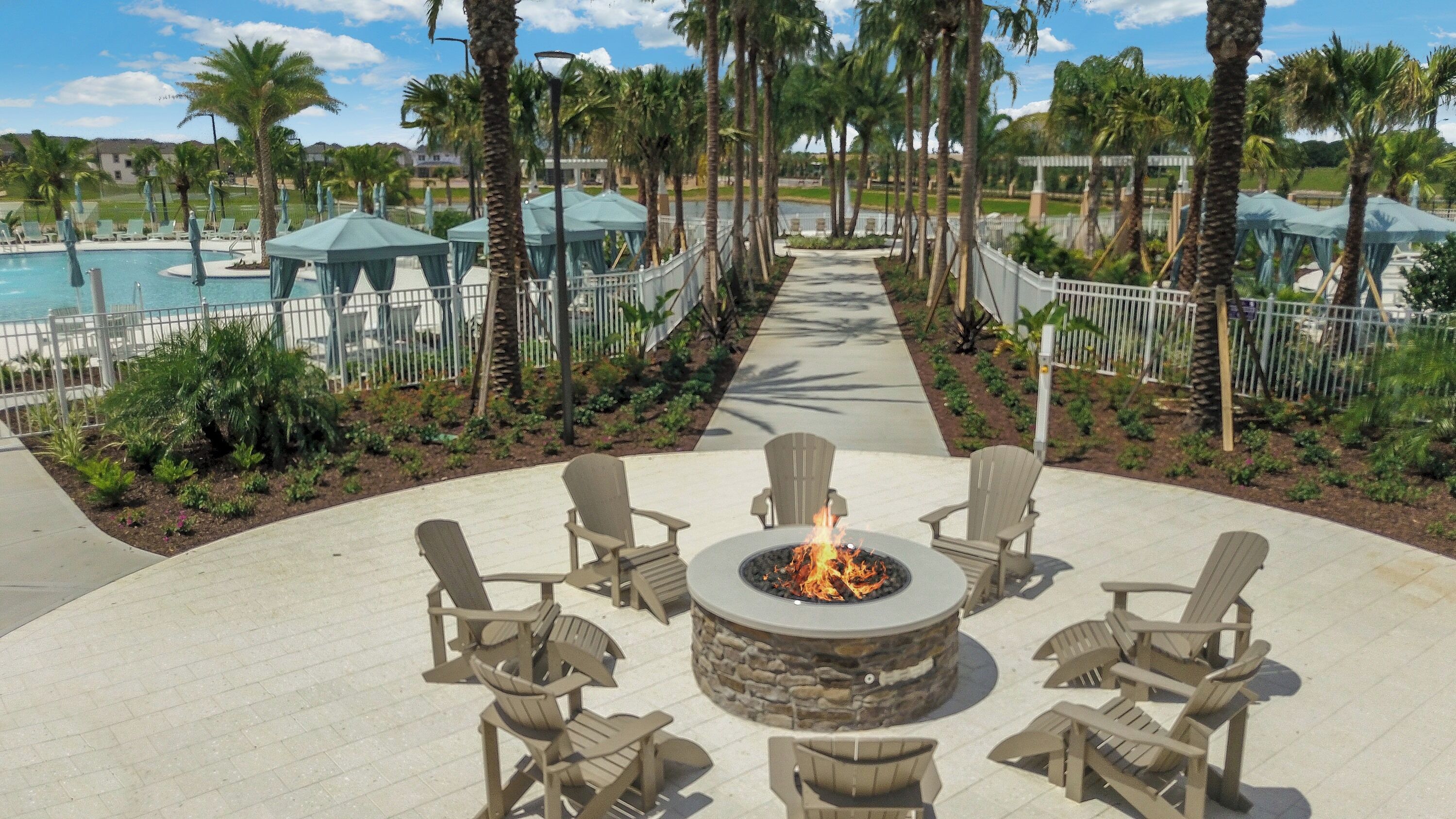 A circular outdoor seating area with a fire pit surrounded by Adirondack chairs, a pathway leading to a pool, and palm trees.
