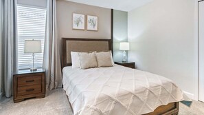 A neatly made bed with a quilt, two bedside lamps, a wooden nightstand, a window with blinds, and two framed botanical prints on the wall. - New Listing Sep 2026/Games Room/Cinema/Themed Bedrooms/Pool&Hot Tub (Kissimmee)