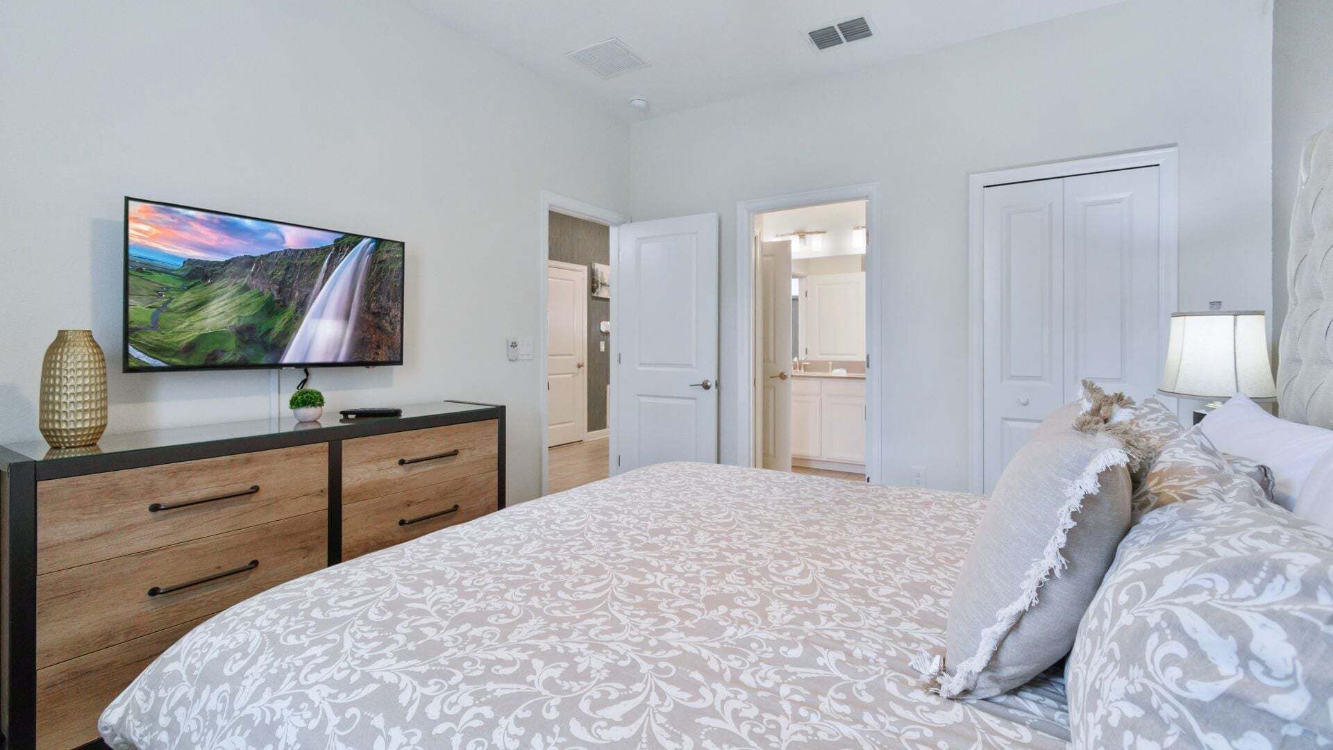 A bedroom with a TV, a bed with patterned bedding, a dresser, and a door leading to a bathroom.
