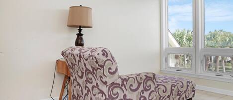 A room with a patterned armchair, a lamp, and a window with a view of palm trees.