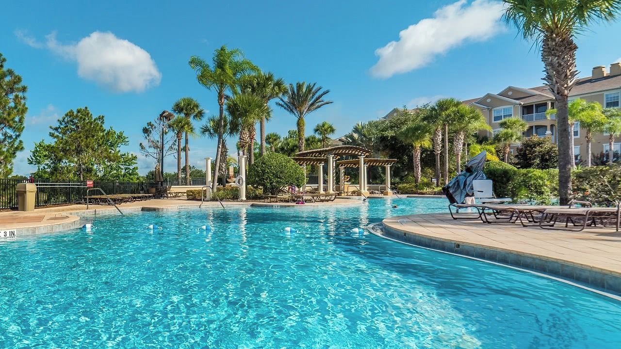 A clear blue pool with lounge chairs and a gazebo, surrounded by palm trees and a building with balconies.