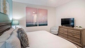 Un dormitorio con una cama, un televisor sobre una cómoda de madera, una silla y dos cuadros colgados en la pared.