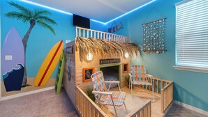 Una sala de juegos infantil con temática playera, que incluye una casita de juegos, tablas de surf y un mural.