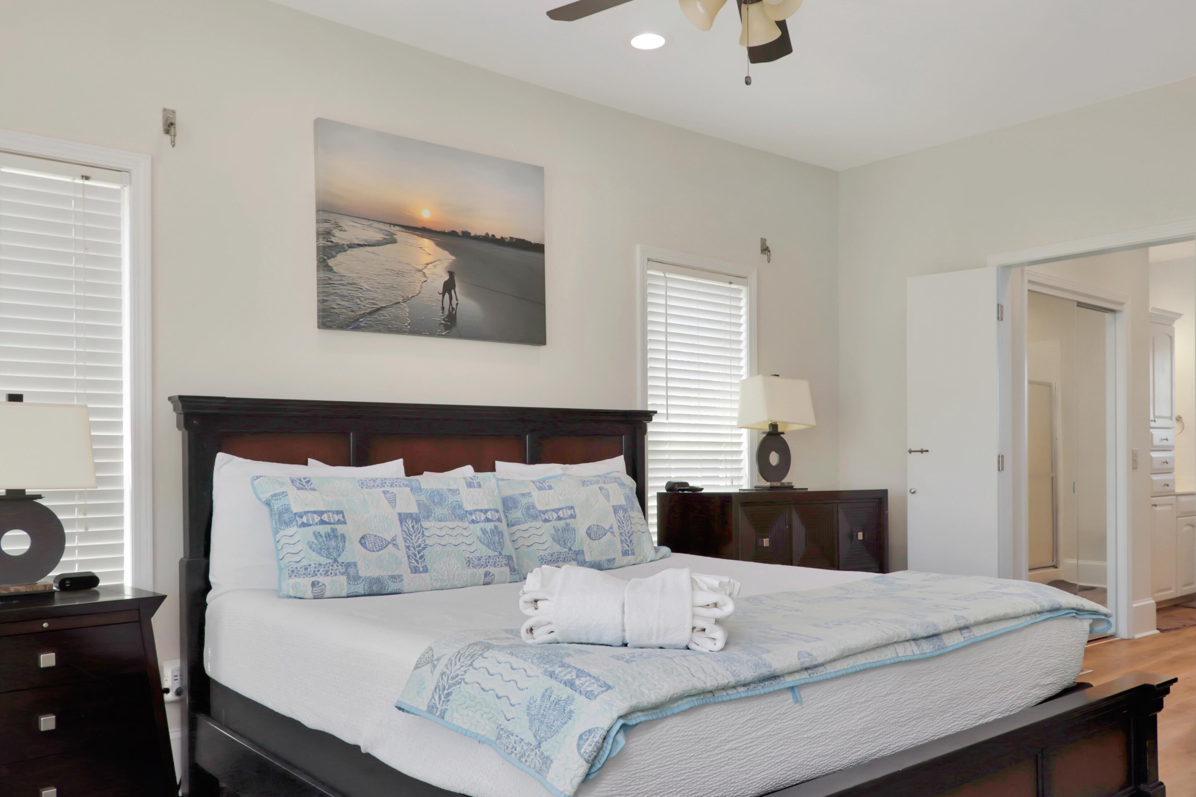 A hotel room with a bed, a nightstand, a lamp, a painting of a beach scene, and a door leading to another room.