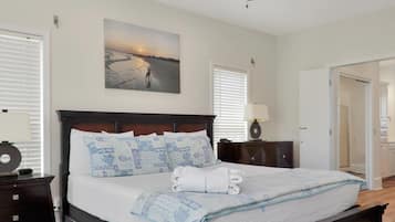 A hotel room with a bed, a nightstand, a lamp, a painting of a beach scene, and a door leading to another room.