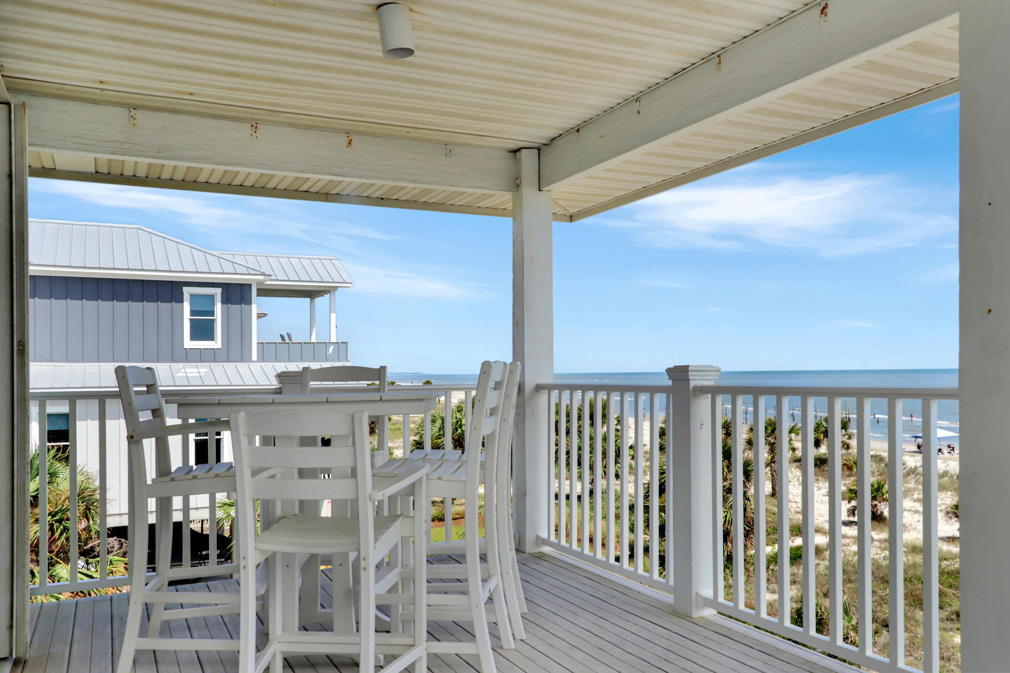 A covered outdoor area with a table and chairs, overlooking a beach and a house.