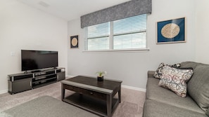Ein Wohnzimmer mit Flachbildfernseher, dunklem Couchtisch, grauem Sofa, gemusterten Kissen und einem Fenster mit Jalousien.