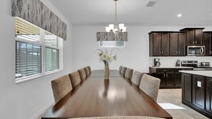 A modern kitchen with dark wood cabinets, a stainless steel microwave, and a dining area with a long wooden table and cushioned chairs. - Mango Shores - Eight Bedroom Villa, Sleeps 16 (Kissimmee)
