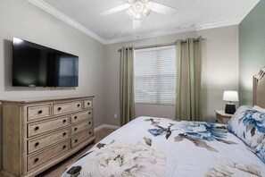 Ein Schlafzimmer mit einem Bett, einem Schminktisch, einem Fernseher, einem Fenster mit Vorhängen und einem Deckenventilator.