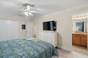 Un dormitorio con cama, ventilador de techo, televisor, cómoda y baño con espejo y lavamanos.