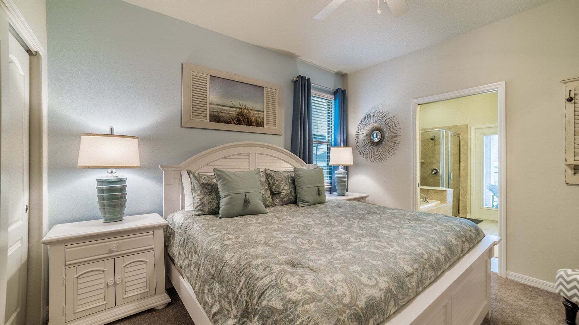 A bedroom with a bed, two bedside tables, a nightstand, a wall-mounted lamp, a window with curtains, a door, and a mirror.