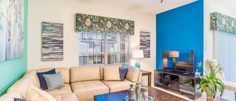 Un salón con un sofá seccional color beige, una mesa de centro con objetos decorativos, un televisor sobre un mueble negro y una pared de acento azul.