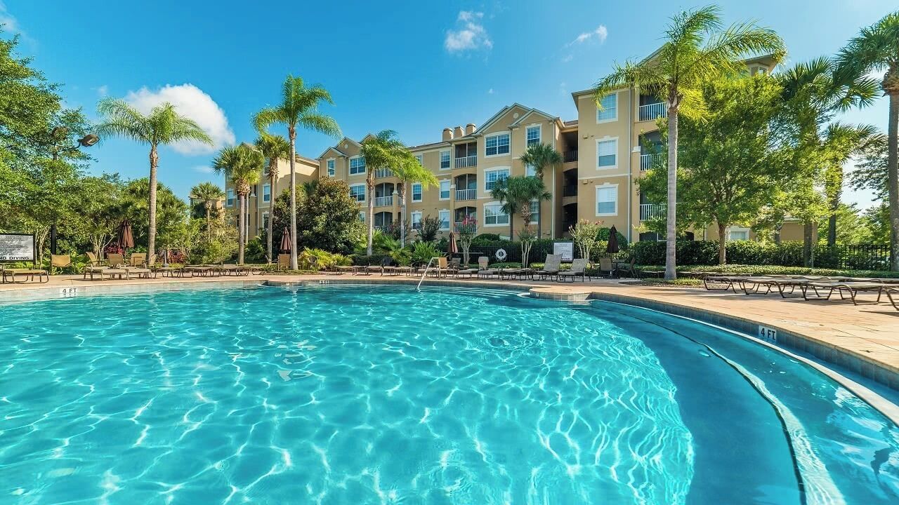 A swimming pool with clear blue water, surrounded by palm trees and a building with multiple balconies.
