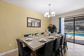 A dining area with a marble table set for four, black chairs, a chandelier, a large mirror, and a view of a swimming pool. - The Grand Slam - Eight Bedroom Apartment, Sleeps 19 (Kissimmee)