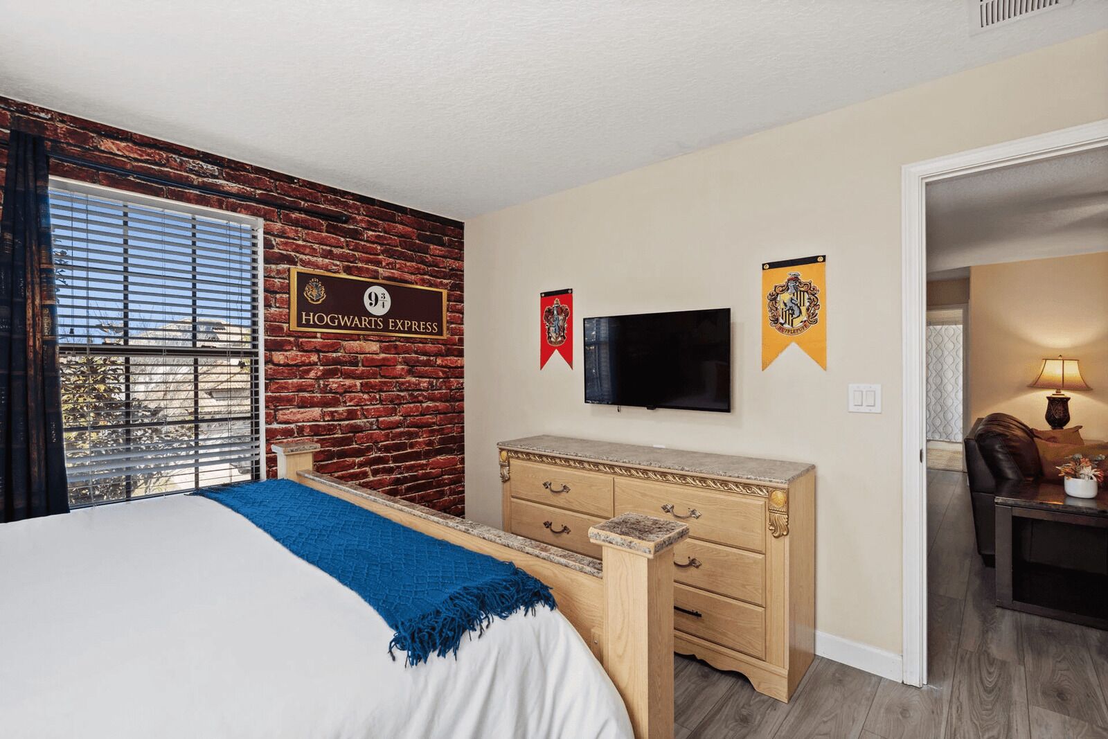 A hotel room with a brick wall, a bed with a blue blanket, a wooden dresser, a flat-screen TV, and two banners.