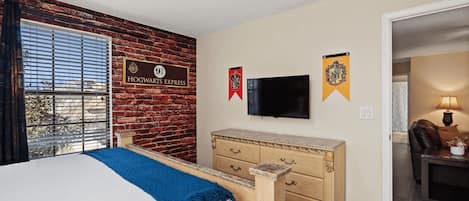 A hotel room with a brick wall, a bed with a blue blanket, a wooden dresser, a flat-screen TV, and two banners.
