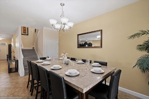 A dining area with a marble table set for four, black chairs, a chandelier, a mirror on the wall, and a staircase leading to another room. - The Grand Slam - Eight Bedroom Apartment, Sleeps 19 (Kissimmee)